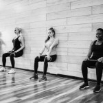 Multiracial group of young athletic slim women doing static exercise with rubber bands leaning on wall of gymnasium in squat